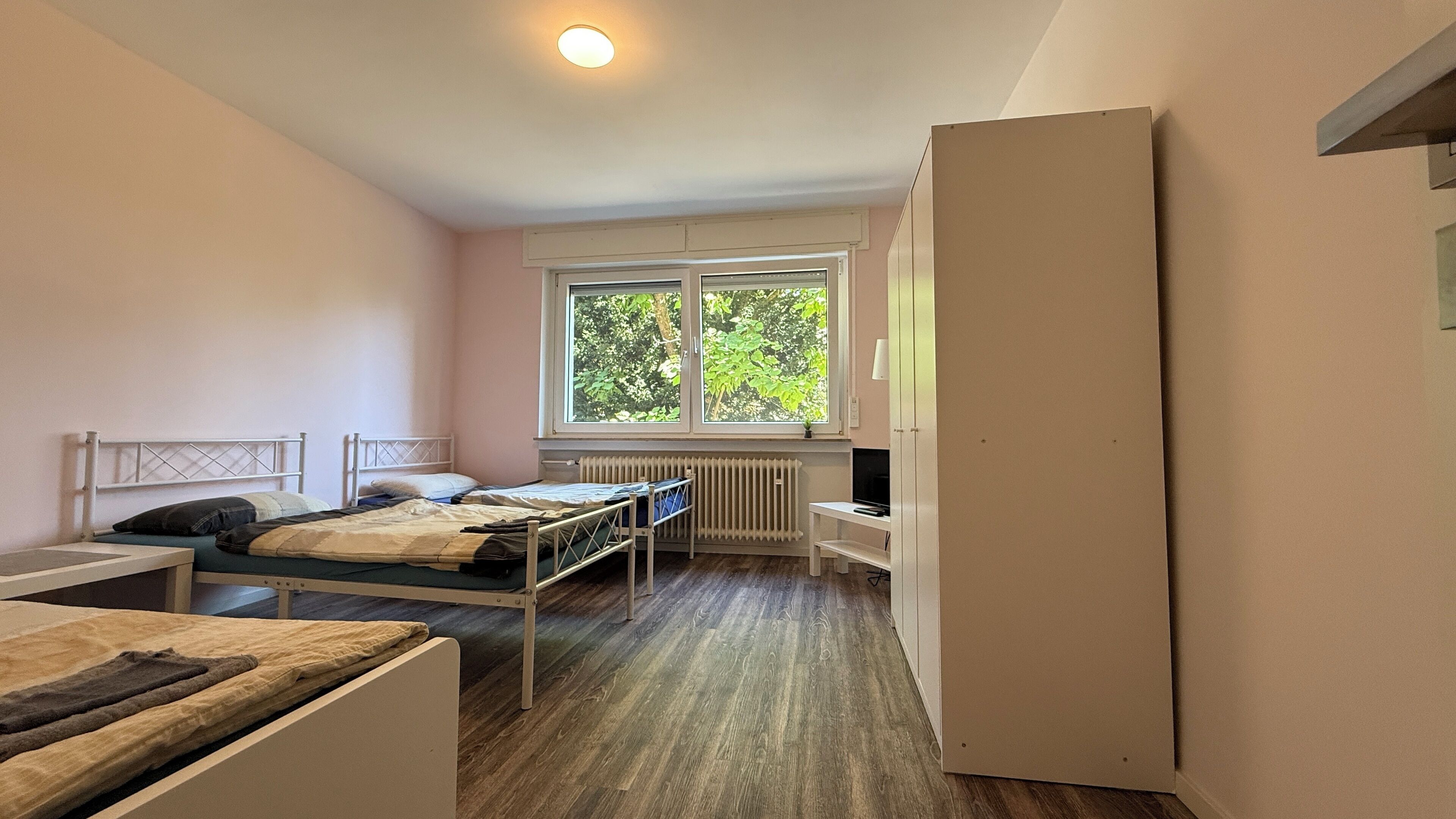 Un dormitorio con dos camas individuales, un radiador, una ventana con vistas a árboles y un armario blanco.