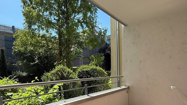 A balcony with a wooden floor and a metal railing, overlooking a green area with trees and a building in the background. - Three Bedroom Comfort Apartment Maintal (Maintal)