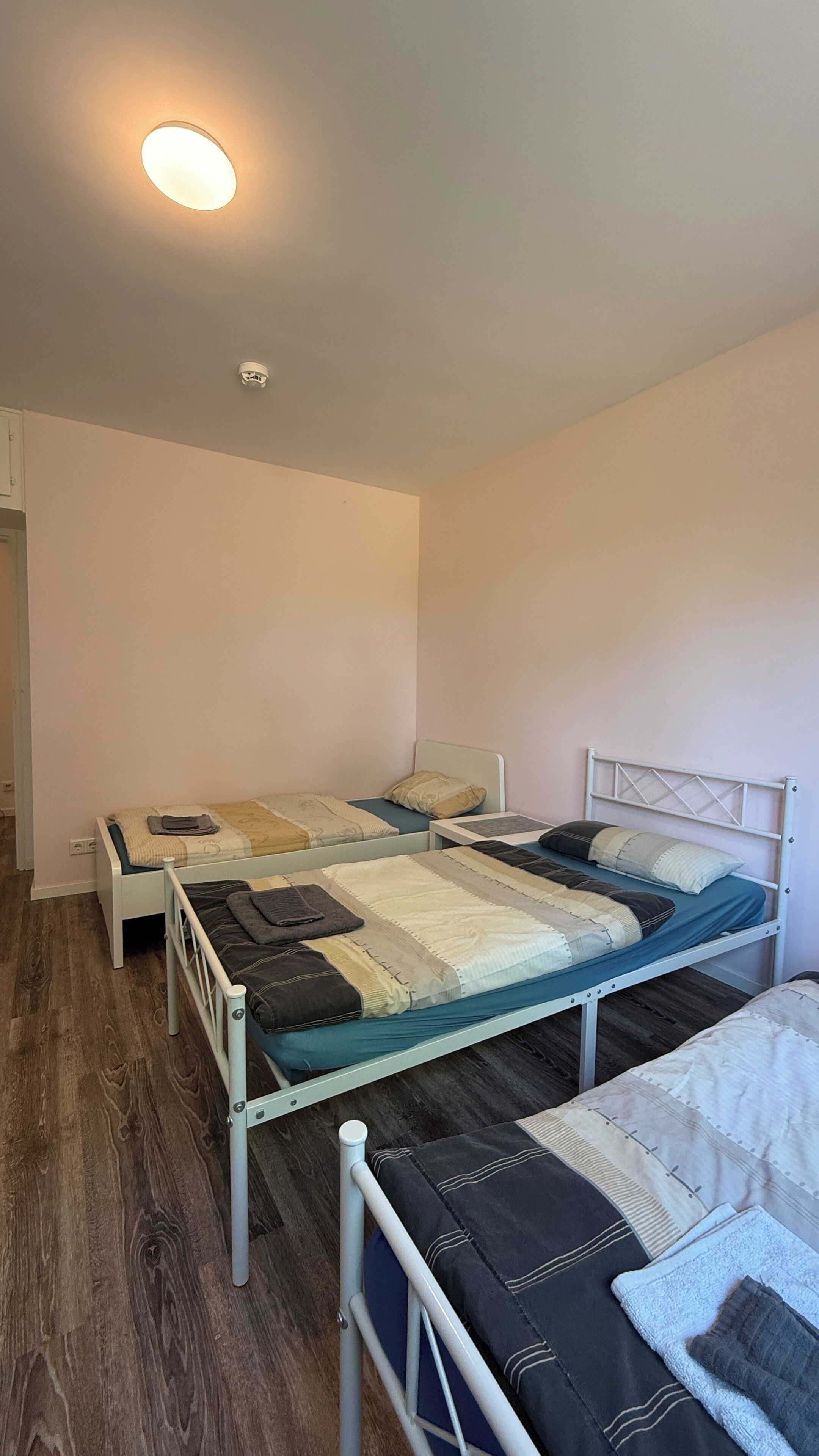 A small, well-lit room with two single beds, each with a different style of bedding. There is a white ceiling light and a smoke detector.