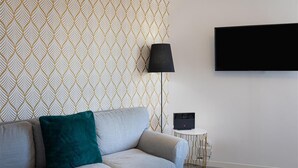 A modern living room with a patterned wallpaper, a sofa with a teal pillow, a floor lamp, a TV mounted on the wall, and a small side table with a speaker. - Vatican Secret Apartment (Rome)