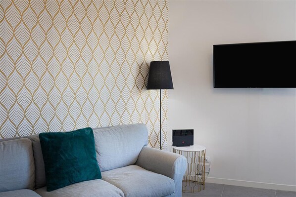 A modern living room with a patterned wallpaper, a sofa with a teal pillow, a floor lamp, a TV mounted on the wall, and a small side table with a speaker. - Vatican Secret Apartment (Rome)
