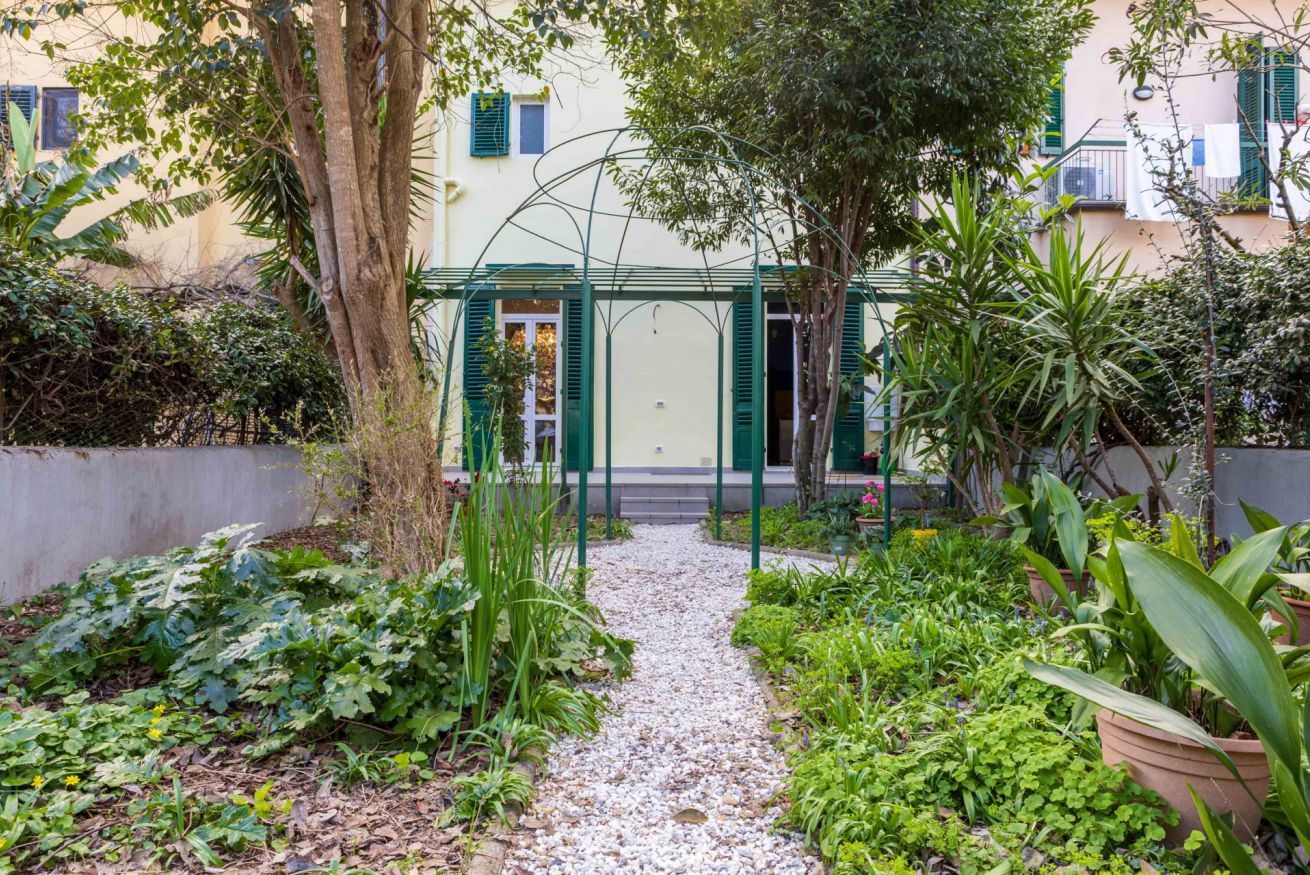 Ein Gartenweg führt zu einem Gebäude mit grüner Tür und Fensterläden, umgeben von verschiedenen Pflanzen und Bäumen.
