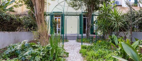 A garden pathway leading to a building with a green door and shutters, surrounded by various plants and trees.