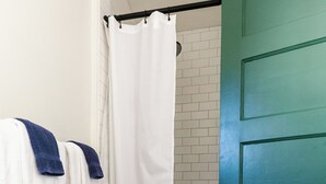 Un baño con puerta verde, paredes cubiertas de baldosas blancas, cortina de ducha blanca y un banco de madera.