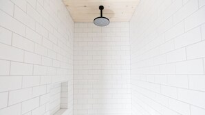 A narrow, white-tiled shower with a wooden ceiling and a black showerhead.