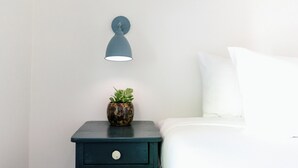 Un lit bien fait, avec du linge de lit blanc, une petite table de chevet dotée d’un tiroir, une lampe fixée au mur et une plante en pot posée sur la table.