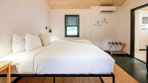 Ein modernes Schlafzimmer mit Holzboden, einem Bett mit weißer Bettwäsche, einem kleinen Fenster und einer Wand-Klimaanlage.