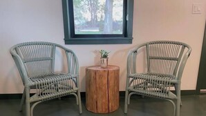 Zwei Korbstühle mit einem kleinen Holztisch dazwischen, ein Fenster mit Blick auf Bäume und ein Lichtschalter an der Wand.