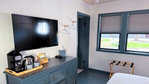 A hotel room with a flat-screen TV, a coffee maker, a bedside table, a bed, a window with a view, and a door leading to another room. - Amber Cottage at Piety Hill Cottages (NEVADA CITY)