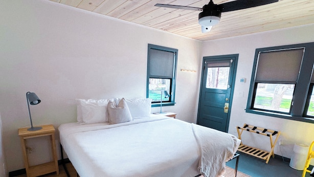 A bedroom with a bed, a bedside table, a lamp, a window, and a door.