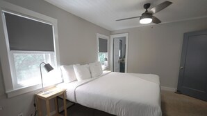 A bedroom with a bed, a desk with a lamp, a ceiling fan, and a window with blinds.