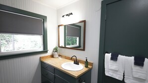 A bathroom with a wooden vanity, a white sink, a mirror, a towel rack, and a window with blinds.