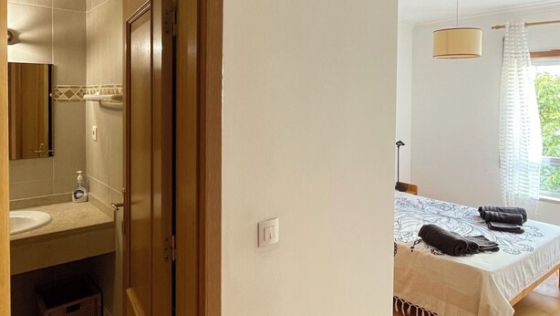 A bedroom with a bed, a bedside table, a window with curtains, and a bathroom with a sink and mirror.