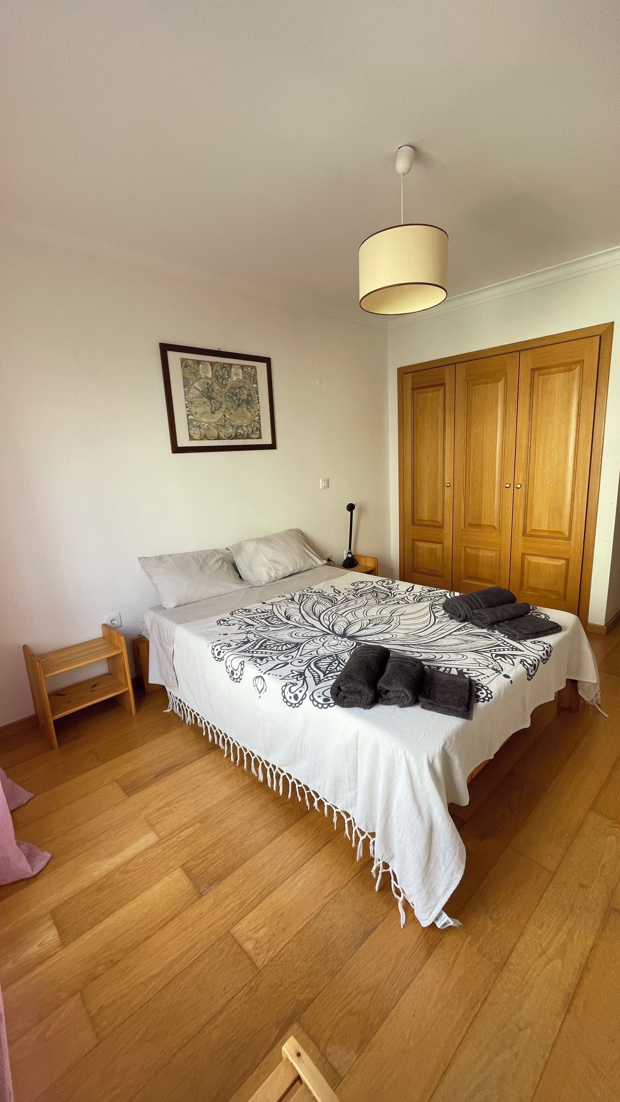 A bedroom with a bed, wooden wardrobe, a small table, a lamp, and a hanging light fixture.