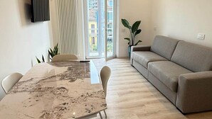 A modern living room with a beige sofa, a dining table with chairs, a large window with a view of buildings, and a radiator on the wall. - Apartment Basilica of Sant'Ambrogio (Milan)