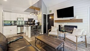 Eine moderne Küche mit weißen Schränken, einer schwarzen Arbeitsplatte und Edelstahlgeräten. Es gibt einen Essbereich mit einem Holztisch und Stühlen, sowie ein Wohnzimmer mit einem braunen Ledersofa und einem Kamin.