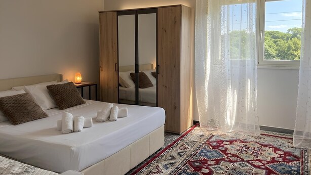 Ein Schlafzimmer mit einem Bett, einem Kleiderschrank, einem Nachttisch, einer Lampe, einem Fenster mit Vorhängen und einem gemusterten Teppich.