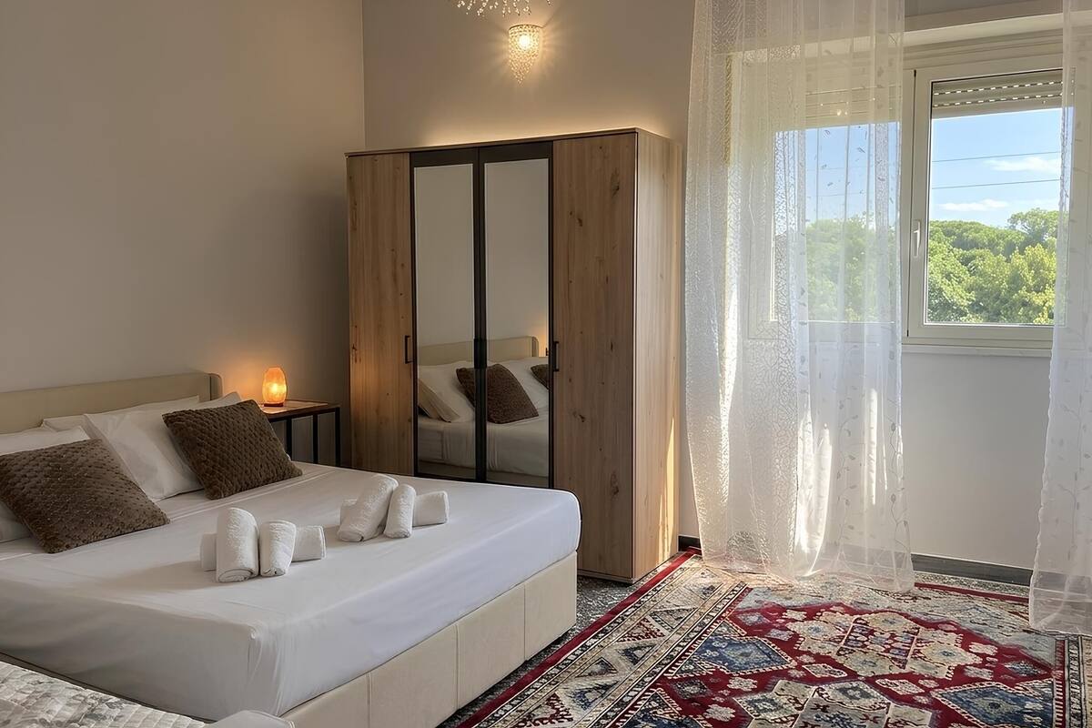A bedroom with a bed, a wardrobe, a nightstand, a lamp, a window with curtains, and a patterned rug.