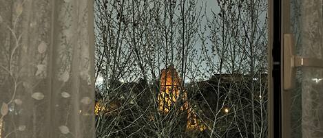 A window view of a building with bare trees in the foreground and a lit structure in the background.