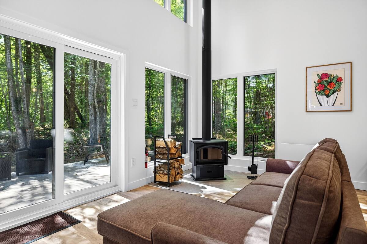 Ein modernes Wohnzimmer mit einem großen Fenster, einem schwarzen Kaminofen, einem braunen Sofa und einem gerahmten Bild an der Wand.