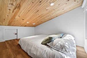 Un dormitorio con techo inclinado, cama con ropa de cama blanca y suelo de madera.