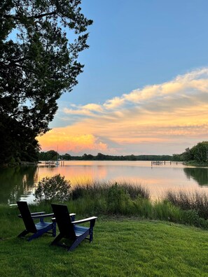 A lakeside view with two Adirondack chairs, calm water, and a sunset.