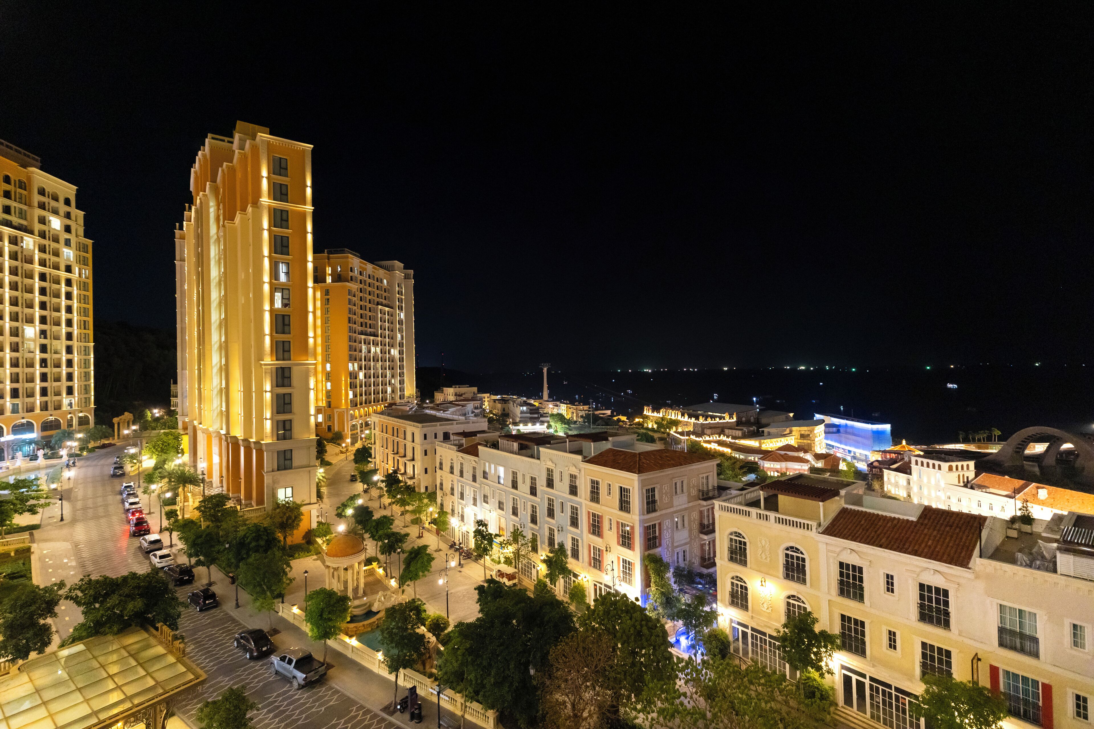 A nighttime view of a cityscape with high-rise buildings, a well-lit street, and a body of water in the distance.