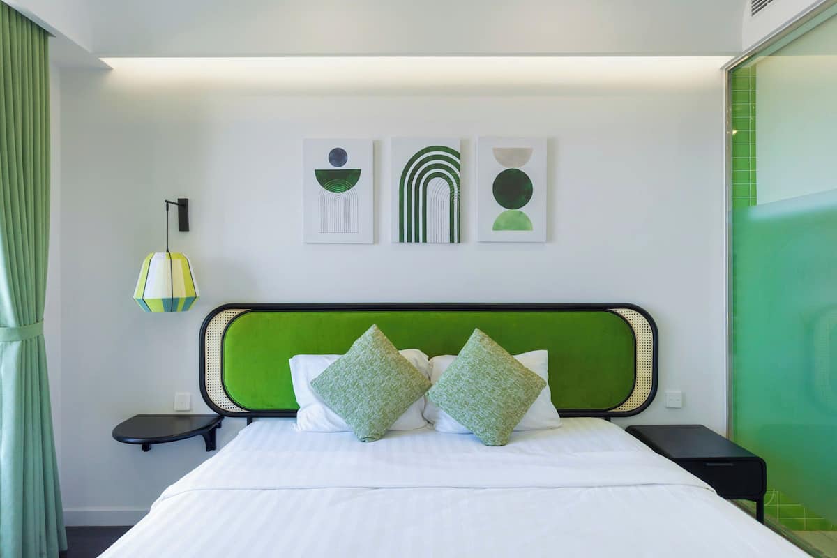 A modern hotel room with a green headboard, a bed with white linens, two green pillows, a bedside table with a lamp, and three abstract wall art pieces.
