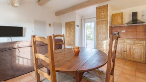 A dining area with a wooden table and chairs, a television in the background, and a kitchen with wooden cabinets and a chimney.