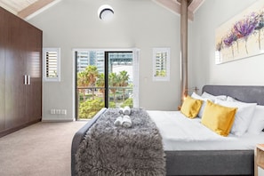 A bedroom with a bed, a grey furry ottoman, a window with blinds, a painting on the wall, and a view of greenery outside.