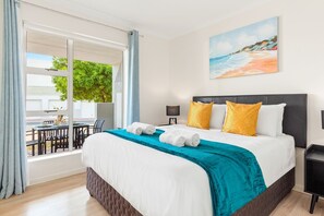 Habitación de hotel con cama, un cuadro de una playa, una mesa con sillas y vistas al exterior.