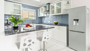 A modern kitchen with white cabinets, a granite countertop, and a built-in oven. There are two white chairs with cut-out backs, and a vase with flowers on the counter. - Big Bay Beach Club 137 (Blouberg)