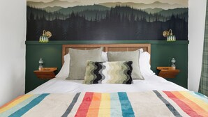 A bed with a rainbow-striped blanket, two pillows, and a headboard with a forest mural. There are two nightstands with lamps and water bottles. - Condo with Backyard Oasis on Ausable River (Wilmington)