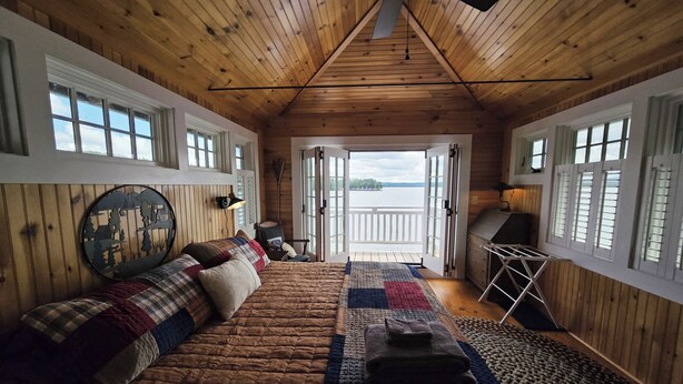 Interior acogedor de una cabaña con techo de madera, una cama grande con colcha colorida, una mesita de noche y vistas a un lago a través de puertas abiertas.