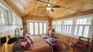 Un dormitorio de cabaña de madera con una cama, ventilador de techo y ventanas amplias.