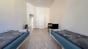 A room with two single beds, a desk with a chair, a wardrobe, and a door leading to another room.