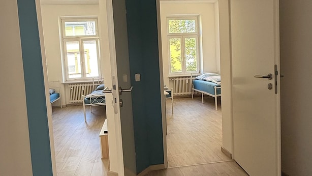 A room with a wooden floor, a door leading to another room, and a window with a view of trees.