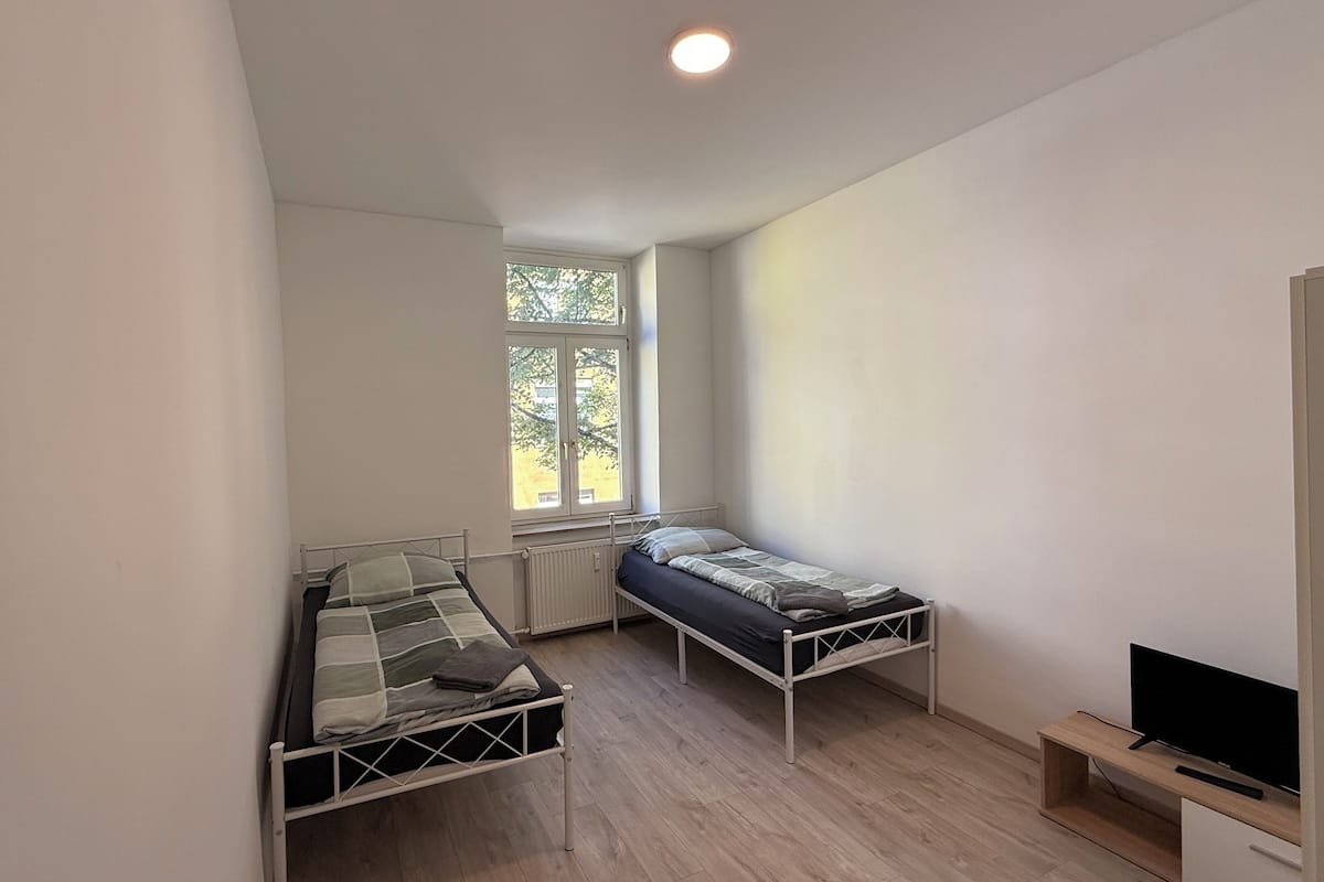 A small, single-room apartment with two bunk beds, a window with a view of trees, a radiator, and a television on a wooden shelf.