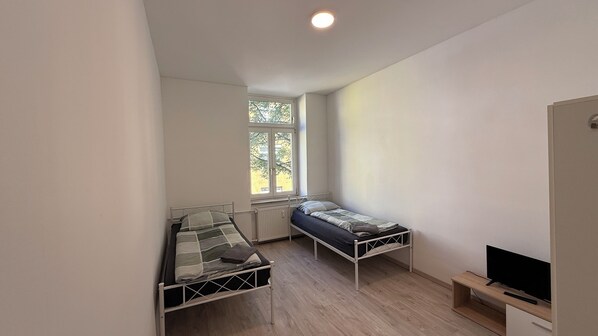 A small, single-room apartment with two bunk beds, a window with a view of trees, a radiator, and a television on a wooden shelf.