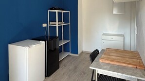 A compact room with a blue wall, a white desk, a black chair, a white refrigerator, and a metal shelving unit.