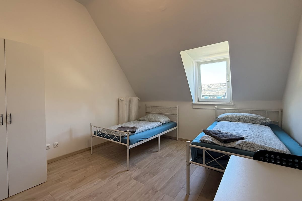 A small, well-lit room with two beds, a wardrobe, and a window providing natural light.