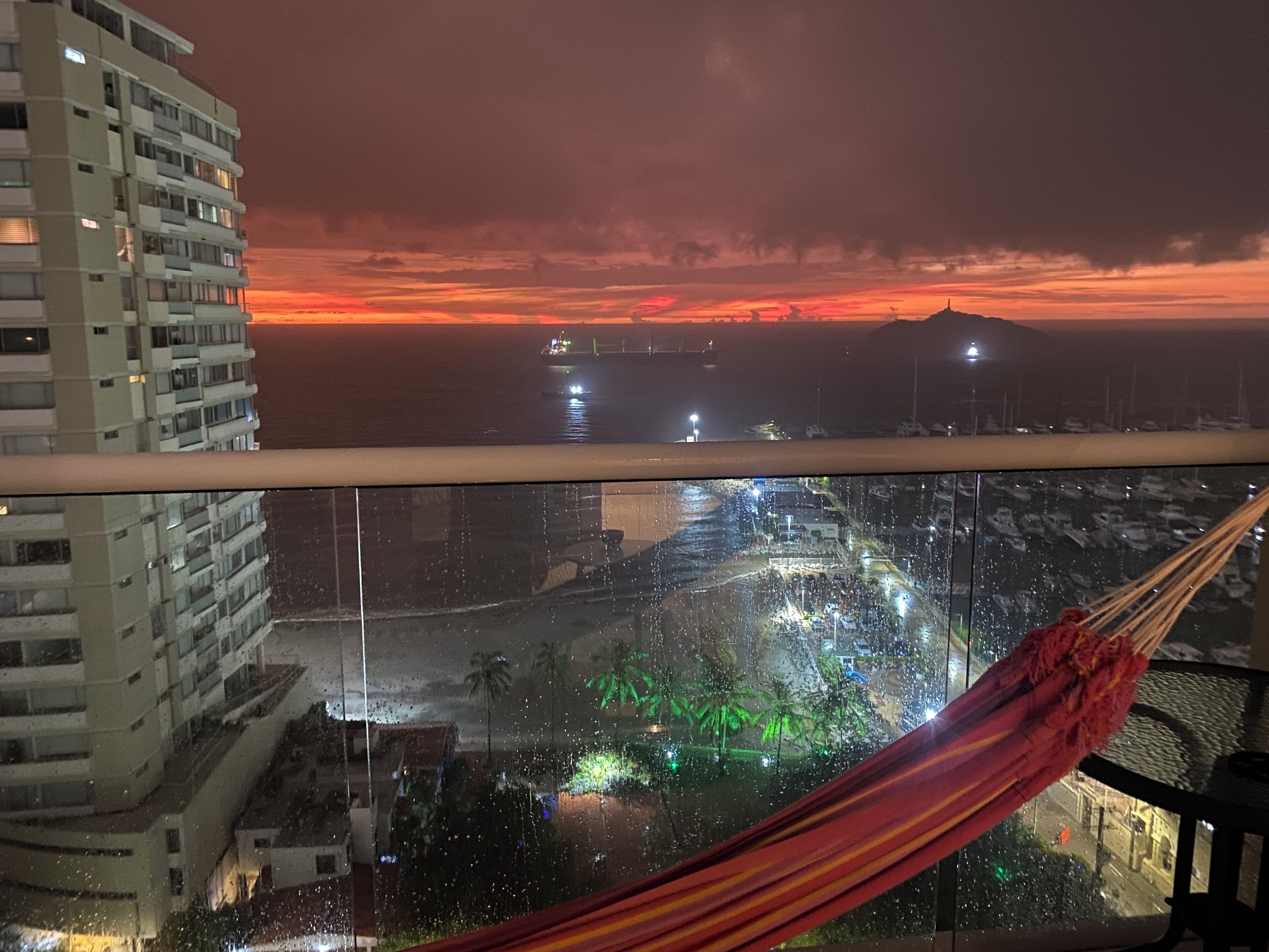 Vista desde un balcón sobre un paisaje urbano nocturno con un atardecer, una hamaca y un edificio de varios pisos.