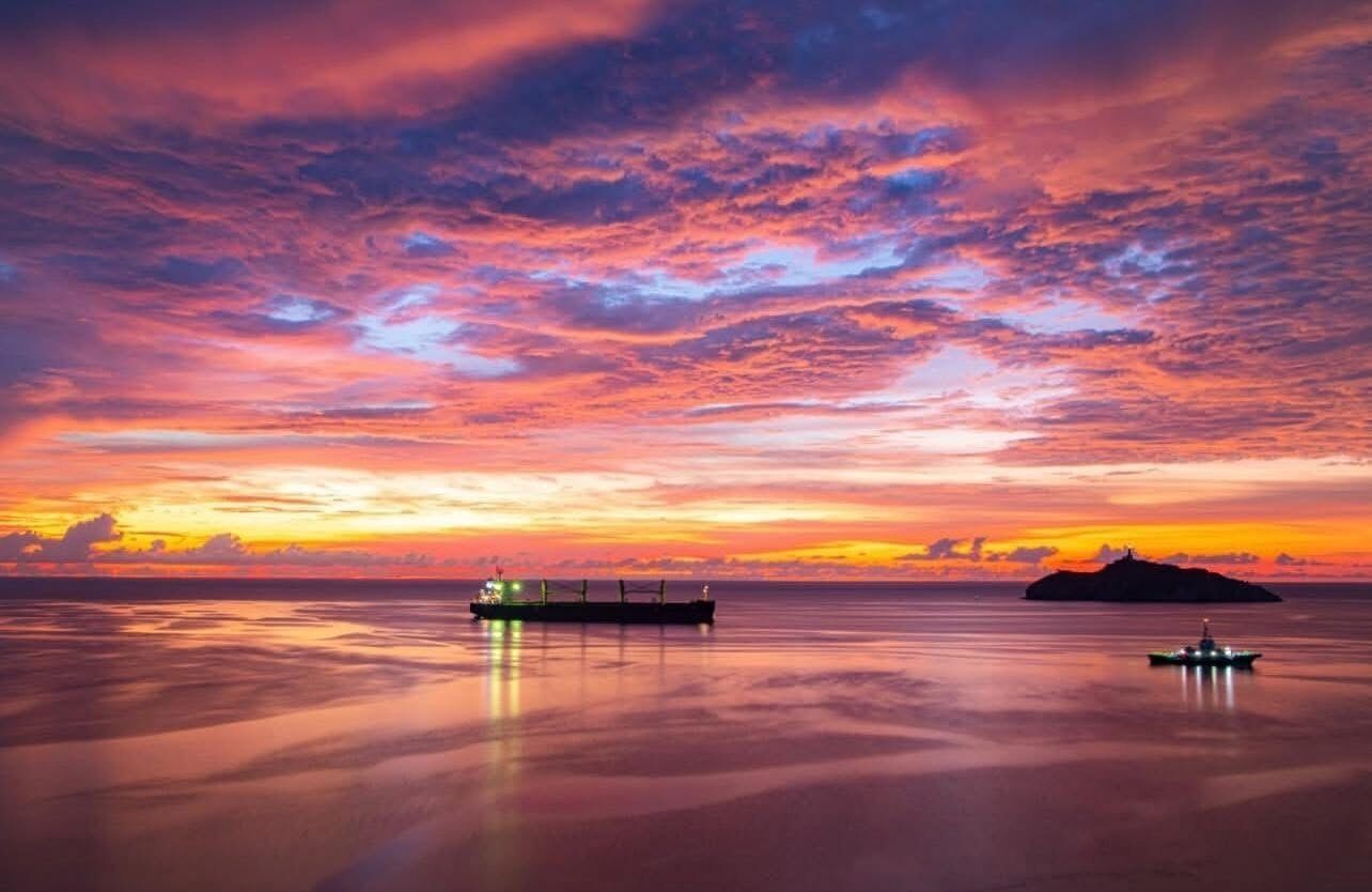 A sunset over calm waters with a cargo ship and a smaller boat.