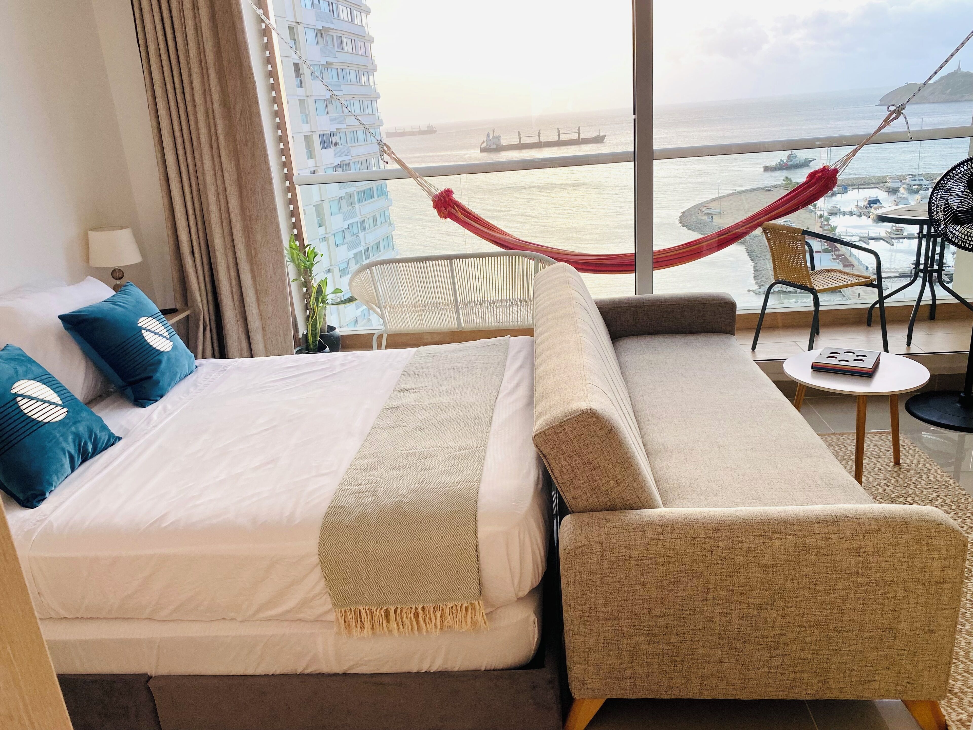 A hotel room with a bed, sofa, and a view of the sea and a bridge.