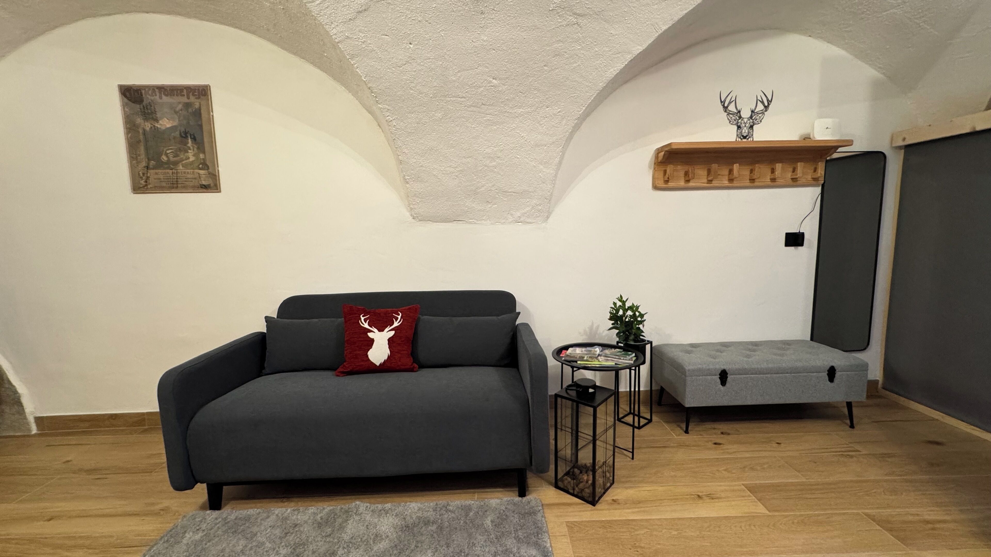 A cozy living room with a grey sofa, a red and white deer pillow, a small side table with a plant, and a grey ottoman.
