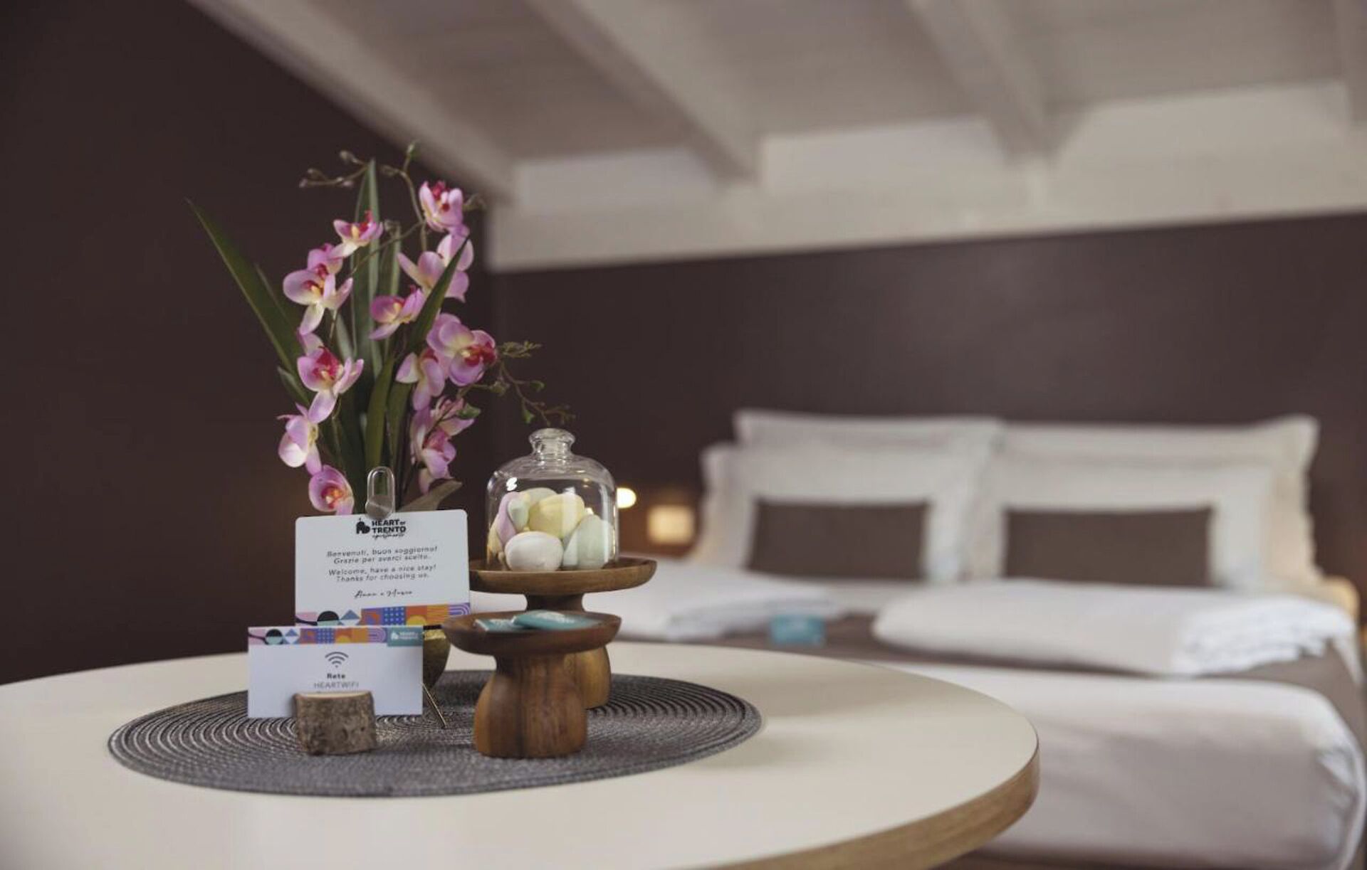 Une chambre d’hôtel avec un lit soigneusement fait, une petite table sur laquelle est posée une carte, un bocal rempli de bougies et un vase de fleurs.