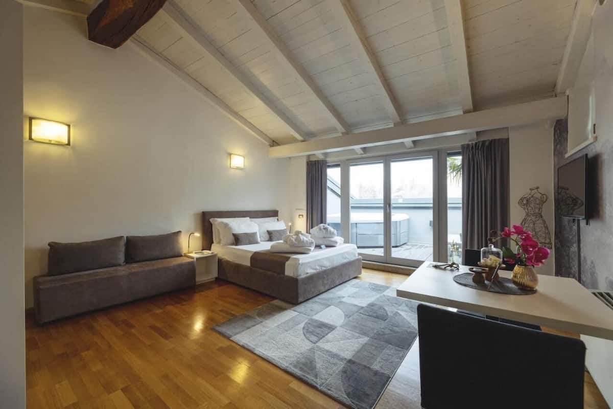 A modern hotel room with a wooden floor, a sofa, a bed with pillows, a desk with a vase of flowers, and a TV on the wall.