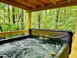 Ein Whirlpool mit zwei Weingläsern, umgeben von einer Holzterrasse und Blick auf einen Wald.