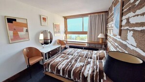 Une chambre d’hôtel moderne, équipée d’un lit, d’un bureau, d’une chaise, d’une grande fenêtre et d’œuvres d’art accrochées aux murs.