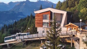 Un bâtiment moderne avec une façade en bois, un balcon en verre et un télésiège en arrière-plan.